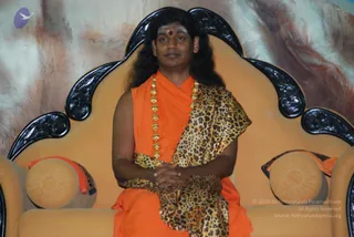 A photograph of a seated man on an ornate ivory-and-black carved throne with cushioned armrests. He wears a saffron-orange robe draped with a leopard-print shawl and a long ceremonial gold bead necklace, and has long dark hair, a red bindi and white vibhuti on his forehead. His hands rest clasped in his lap, and a glossy marbled backdrop fills the background. Several Swamiji cues are present: swept-back long hair, saffron clothing, gold ornaments and a throne seat, so the appearance is a probable match for Swamiji seated in person.