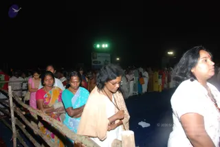 Night photo of a long queue of women and a few children standing behind a wooden railing along a dark dais or open ground. The foreground woman in the centre wears a tan shawl over a white top with a rudraksha-style beaded mala, glasses, hands folded in prayer. Beside her a woman in a teal blouse and multicoloured sari stands with hands clasped, and a woman in a red blouse and yellow-green sari stands near the left. On the right a woman in a white top with a red bindi and beaded necklace looks sideways. Behind them stage lights glow green and a billboard-style banner is visible in the background. No visible murti or ritual object is clearly shown.