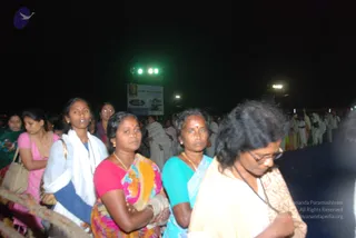 A night-time photo of a crowd queuing along a rope barrier at an outdoor event. In the foreground, a woman in a white shawl and pink-and-white top stands beside a woman in a red blouse and multicoloured sari, and a woman in a teal blouse with a yellow sari and a red bindi. At the far right, a woman in glasses and a cream sari with a dark beaded necklace looks down. Behind them, people in white ritual clothes stand in a line, and a large banner on a stand is visible beneath bright lights. No a person candidate is clearly identifiable in the scene.