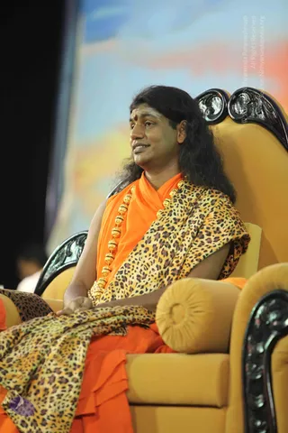 A man with long dark swept-back hair sits on an ornate yellow throne-style armchair with carved black wooden armrests and backrest. He wears an orange saffron robe, a gold bead necklace of large round beads, and a leopard-print shawl draped over his shoulders and lap. A red bindi marks his forehead. Behind him is a large blurred stage backdrop in blue, orange and pink tones. Swamiji (probable) is seated on the throne, wearing saffron robes, a gold bead necklace and a leopard-print shawl, with long swept-back hair and a bindi, in what appears to be a stage setting with a backdrop.