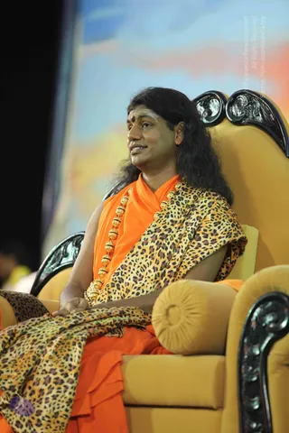 A man with long dark hair and a red bindi sits on an ornate golden throne-style chair with carved black wooden armrests against a blue and orange backdrop. He wears orange robes, a long gold bead necklace, and a leopard-print shawl draped over his shoulders and lap. He appears to be seated on a stage during a talk.