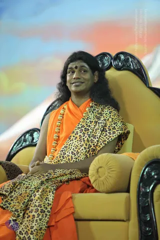 A seated man with long, swept-back dark hair sits in an ornate yellow throne with dark carved wooden arms and a round bolster cushion. He wears a saffron-orange robe draped over a leopard-print shawl and a long gold bead necklace, with a red bindi on his forehead. He sits with his hands clasped in his lap and looks off to the side with a slight smile. Behind him is a soft painted backdrop in blue, orange and pink. Visible cues in the appearance and attire are consistent with a person seated in a position of honour, identified here by appearance only.