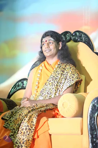 A photo of a seated man in saffron-orange robes draped with a leopard-print shawl, resting his hands clasped in his lap on an ornate yellow throne with black carved armrests. He has long swept-back dark hair, a red bindi on his forehead, white ash markings, and wears a long beaded gold-toned necklace. A yellow cushion rests on the armrest and a colourful blurred backdrop fills the background. Swamiji (probable): the long hair, saffron clothing, beaded ornate necklace and throne seating are the qualifying cues for this figure.