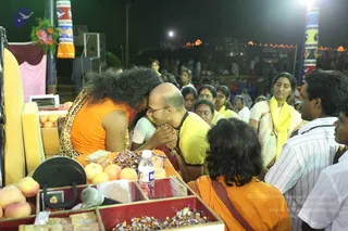 Night outdoor darshan scene. A person with long, wild dark hair wearing an orange sleeveless top and leopard-print wrap leans forward over a yellow-cushioned seat to greet a bald man in glasses and a yellow shirt, who bows with hands pressed together. Before them is a table with apples, fruit, wrapped sweets, a water bottle, a microphone and open wooden boxes. Around them stand devotees: a woman in a white sari with a yellow shawl, another in yellow, and men in white shirts. Strings of lights and colourful painted pillars frame the stage behind them.