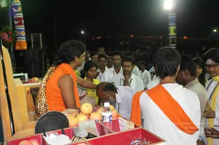 Night-time outdoor event photo. A man with long swept-back dark hair wears an orange sleeveless top with a leopard-print shawl and stands at a wooden table covered with apples, a water bottle, a microphone and red-lined trays. He reaches out to touch the head of an older bearded man in white who bows with hands folded. Around them, men in white shirts and women in yellow garments stand watching; one young man wears an orange sash and rudraksha-style beads. Multi-coloured decorated poles and string lights frame the background crowd.