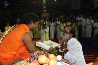 A photograph of an indoor event at night. On the left, a man with long dark hair swept back wears a saffron-orange sleeveless garment and a leopard-print shawl over his shoulder, seated on a raised platform. He holds out a white cloth bag embroidered with a gold motif toward an older man on the right, who has a white beard, glasses, a gold wristwatch and a ring, wears white clothing, and sits with palms pressed together in greeting. In front of them lie apples, wrapped chocolate candies and a water bottle on a cloth. Behind them, a crowd of devotees stands in white and yellow garments, and a tall decorated pillar with orange and white banding rises in the background.