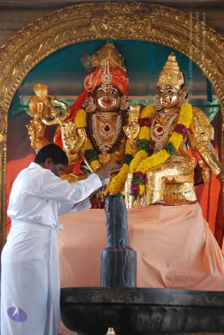 A photograph of a Shiva lingam being bathed during an abhishekam at a shrine. A man in a white dhoti and white shirt stands at left, pouring liquid from a vessel over a dark black stone lingam set on a black base. Behind it, two gilded deity murtis sit under a gold arched frame: the left murti wears a red turban with a jewelled crown and a rudraksha mala, holding a trident, while the right murti wears a golden crown and jewelled necklaces. Yellow and magenta marigold garlands drape both figures, and an orange-peach cloth covers the platform. No living a person is visible; the figures are statues.