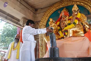A photograph of a temple abhishekam at a shivalinga. A man in white dhoti and shirt stands at center, pouring liquid from a metal vessel onto a black stone shivalinga set on a dark base. Behind him stands a deity murti in gold, with a red and gold crown, red sash, gold ornaments and a large garland of yellow and pink flowers, seated on a peach-coloured cloth. To the left, a bearded man in white robes with a yellow shawl and a beaded necklace watches the ritual. The setting is a stone temple pavilion with carved gold-framed arch, a green backdrop and foliage visible outside.