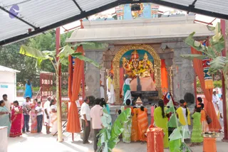 A photograph of an open-sided temple shrine under a corrugated roof, with orange cloth drapes and banana-leaf decorations framing the arch. Inside the golden arch stands a deity murti with a crown, flanked by two smaller figures, garlanded with yellow and orange flowers. A priest in white dhoti and shawl stands beside a black stone shivalinga-style basin being bathed, as in abhishekam. Devotees in saffron, orange, green, pink and blue saris and white dhotis stand in a queue at the left and right, some holding offerings. A small brass lamp and kalasha-like vessel sit at the front. Tile floor and trees appear beyond the railing.