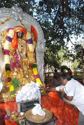 Outdoor shrine photo of a gilded Dakshinamurthy murti seated under a silver arch and a large tree. The golden deity wears an orange turban-like cloth, a rudraksha-style mala and a heavy necklace, and is garlanded with white, yellow, pink and purple flowers. In front, white cloth bundles are piled on a stone base with a copper plate holding fruit and offerings. A man in a white shirt and white trousers kneels at the right, one hand touching the offerings. Two or three figures stand in the background among the trees. The orange-painted rock base is visible in the foreground.