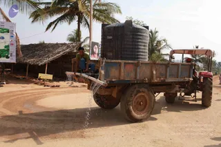 A tractor pulling a rusty blue-and-red flatbed trailer carries a large black water storage tank labelled Reno. A young man in a green shirt and blue jeans sits on the trailer's pipe, with water spraying from a pipe outlet onto the dirt road. Behind is a thatched-roof shelter with a yellow sign at its base, a banner with a green map and a portrait of a woman in a pink top, and tall palm trees under a pale sky. This appears to be a water supply or distribution scene at a rural ashram site.