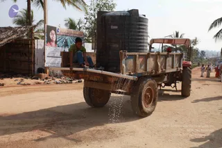 A dusty dirt road in a rural village with palm trees and a thatched hut at left. A young man in a green shirt and blue jeans sits on the rear edge of a red tractor's flatbed trailer, working a tap so water streams from a pipe onto the road. A large black water storage tank labelled Reno sits on the trailer. Several people in white and light clothing walk along the road in the background. A roadside banner at left shows a smiling woman's portrait with printed text and a small temple illustration, and a row of sandals lies on the ground near the hut.