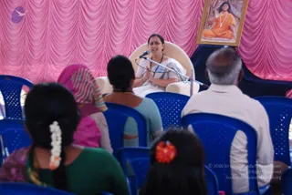 A woman in a white sari with a light border sits in a cream armchair on a stage draped with pink swagged curtains, speaking into a microphone on a stand while her hands are held together. Behind her hangs a framed portrait of a figure with long dark hair in saffron/orange robes and a beaded necklace, seated in meditation before a tree. Several seated audience members in blue plastic chairs face the stage: a woman with a white flower in her hair in green, a woman in a striped headscarf, a woman in teal, a girl with a red flower hairpin, and an older man in a white shirt. The scene looks like a talk or satsang.