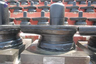 Close-up photograph of a row of polished black stone Shiva lingams standing on carved yoni-style pedestals against a red-painted brick wall. The foreground lingam sits on a square grey stone base with a small metal plaque reading installed by Raman, Lalitha, Jayashree, Sriram and Jayaram, Texas. No people are visible in the scene.