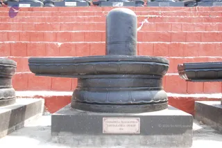 A photograph of a black stone Shiva lingam on a yoni-shaped base, displayed outdoors in front of a tiered red brick wall. Several other black stone lingams stand on plinths along the upper steps, and a strip of coloured string or flags hangs across the top of the wall. No people are visible.