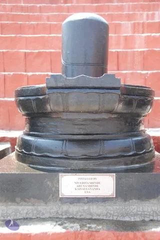 A large dark polished stone Shiva lingam stands on a multi-tiered black yoni-pitha base set on a grey plinth against a red brick wall. A brass-coloured plaque on the plinth is inscribed with the installers' names.