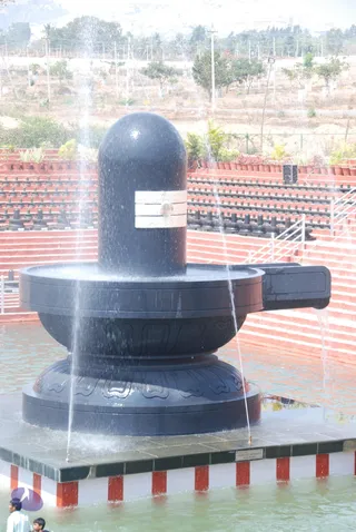 A large black stone Shiva lingam with a yoni-pitha base stands in a shallow pool, with water streaming down its surface from overhead jets. The lingam has a white decorative band across its upper section. Terraced red-brick steps lined with small black lingams rise behind it, with potted plants and trees beyond and a dry hillside in the distance. At the lower left edge, two partly visible people, including a child, stand near the pool edge. A small plaque sits on the base platform.