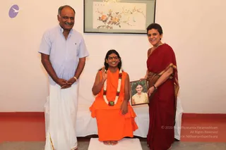 Three people stand and sit in a white-walled room under a framed floral painting of birds and blossoms. A man on the left wears a light blue short-sleeved shirt, white dhoti and a gold-toned wristwatch, hands folded. In the centre, a person sits cross-legged on a low white-draped platform in saffron-orange robes, wearing a white and red flower garland, a red bindi and a waving raised right hand, appearing as a probable Swamiji by visible cues of long swept-back hair and saffron clothing. On the right, a woman in a deep maroon sari with gold border holds a framed photograph of a young woman in a yellow outfit.