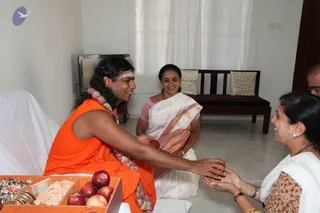 A photograph of a home visit in a plain white room with a wooden bench and cushions behind. A long-haired man in an orange sleeveless garment with a white tilak on his forehead and a pink-and-white flower garland sits on a white sheet and reaches out to hand a red apple to a woman kneeling at right in a white sari with a brown border. Another woman in a white sari with gold border sits smiling in the middle, and a partly visible man stands at the top right edge. A wooden tray in the foreground holds apples, peaches, sweets, and small packets. The man has distinctive long swept hair, orange robes and a garland, so this is a probable appearance-based candidate for a person seated in person.
