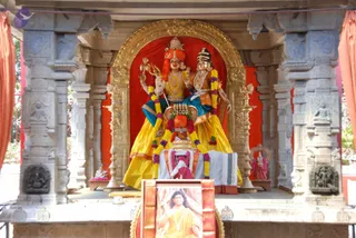 A photograph of an outdoor shrine with a large deity murti pair seated on a raised pedestal. Two painted figures stand side by side: the left one has a red and orange crown, holds a trident, and wears gold necklaces and yellow dhoti; the right one wears a crown with a blue-and-orange garment and gold ornaments. Yellow and orange cloth drapes the pedestal, garlanded with yellow and purple flower garlands, and a kalasha sits in front. Ornate gold arch frames the shrine, with grey stone pillars carved with figures and brass oil lamps standing to each side. A small framed portrait sits at the base, showing a man in saffron robes with a mala, though the photo gives no clear view of his face. No people are visible in the scene.