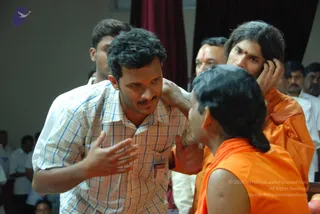 A photograph of a dimly lit hall where a man in a checked short-sleeved shirt with a visitor badge stands gesturing with open hands in conversation. A young sannyasi in orange robes with long swept-back hair and a red bindi holds his head with both hands, and another orange-clad monk with a tied-back ponytail stands in the foreground. Behind them men in white shirts and a red curtain are visible.