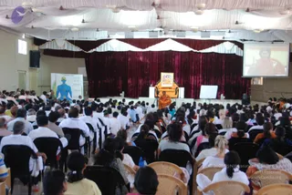 A large hall viewed from the back, filled with rows of seated attendees on black plastic chairs, mostly in white and light clothing. On a raised stage at the far end, a figure in orange robes sits on an ornate golden throne-like chair beneath a framed portrait, with a magenta velvet drape behind. A large screen on the right shows a close-up of the same orange-robed figure. To the left, a standing chart shows a blue human figure labelled with chakras. White drapes hang from the ceiling and a Chakra chart stands at the side. a person: the seated figure on the throne wears orange robes and long hair, consistent with a presiding guru, while the attendees are devotees seated in rows.