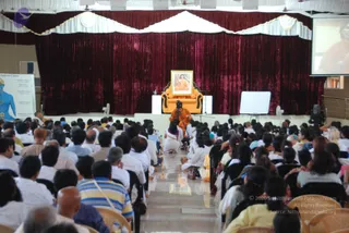 A large hall audience seated on white chairs and floor cushions faces a stage with a maroon velvet curtain and white valance. On the stage, an orange-upholstered throne-like chair sits beneath a framed portrait of a figure in orange robes, and a seated figure in orange clothing sits near a camera on a tripod. A whiteboard and a chakras chart on the left wall are visible, and a projection screen on the right shows a close-up of a face. Many people in white, yellow and striped shirts fill the rows in the foreground.