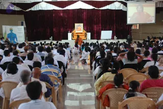 Wide view of a large indoor hall during a seated satsang or a person Spurana Program session. Hundreds of seated attendees, mostly in white and light clothing, face a raised platform. On the platform a figure in orange robes sits on an ornate orange throne in front of a framed portrait showing a figure in orange. A maroon velvet drape with white scalloped trim hangs behind the stage. A large screen at upper right shows a close-up of the speaker in orange. At left, a chakra chart displays a blue human figure with seven chakras, and a whiteboard stands beside the stage. Some attendees wear orange, pink and yellow saris, and a few sit with hands raised.