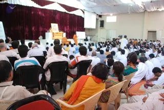 A wide photograph of a large hall during a seated convention session. A crowd of attendees sits on rows of plastic chairs, many in white clothes, some in orange and yellow saris and shawls, several wearing headphones. At the front, a raised stage with maroon curtains holds a golden throne-like seat with a framed portrait on a stand. A standing figure in orange robes stands near a whiteboard on the stage, and a cameraman in white stands at the right edge. A projection screen hangs at the top. The people count is approximate and large.