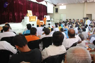 A photograph from behind a large seated audience in a hall at a convention session. On the raised stage at left, an orange velvet-upholstered throne sits empty before a maroon velvet drape, with a framed portrait placed on a white pedestal beside it. A figure in an orange robe stands near a white flipchart at centre stage, and a projector screen is mounted on the back wall. Audience members in white shirts, cream and orange clothing sit on black chairs and floor mats, many with shaved or grey heads. A cameraman stands at the right edge, and a figure in white with an orange sash stands near the right side.