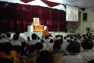 A wide photograph of an indoor hall during a seated session. On a raised white platform stands an orange-upholstered armchair in front of deep magenta velvet drapes, and a gilded framed portrait of a saffron-robed figure sits on a small stand beside it. A figure in saffron/orange robes with long hair sits in the chair, holding papers or a book. A projection screen at upper right shows a close-up of the same saffron-clad figure speaking into a microphone. In the foreground, a large audience in mostly white clothes sits in plastic chairs, several wearing headphones, with a video camera on a tripod at right.
