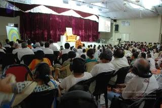 A photograph taken from the back of a seated audience in a large hall during a session. Rows of attendees in white and pastel clothes sit on black plastic chairs, many wearing over-ear headphones for translation. On a raised stage draped with maroon velvet curtains, a figure in orange robes sits on an orange cushioned chair before a framed portrait on a white pedestal. A projection screen on the right shows a image, and a colourful concentric circle graphic stands on the left.
