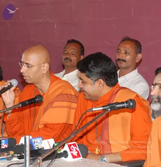 A press interview scene with saffron-robed sannyasis seated at a table covered in microphones and news channel logos including Udayan, Sun, ETV and Star. A bald man in orange robes and glasses holds a microphone and speaks, while the central seated man in orange, with a rudraksha mala and a gold pendant, smiles beside him. Two men in white shirts stand behind them against a maroon wall, and a bearded man in orange appears at the right edge. The seated man in the centre has no distinctive crown or swept hair cues, so his identity remains uncertain.