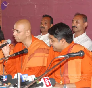 Photograph of a press interview at a press conference table with multiple microphones and news channel logos visible at the front. Two men in saffron-orange robes sit at the table: the man on the left, bald with glasses, gestures with one hand while speaking into a microphone and wears a red bindi and a long beaded necklace; the man on the right, with dark swept hair, listens with a downward gaze and wears a saffron garment and a pendant on a cord. Behind them stand two men in white shirts against a maroon wall.