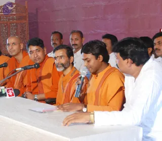 A press conference or interview at a table draped in white cloth, with several microphones and a red-and-white channel logo on the table. A man in an orange kurta with a shawl holds a handheld microphone and speaks while seated. Beside him, a bearded man in orange robes with rudraksha beads and gold-rimmed glasses sits attentively. Further left, a shaven-headed man in orange with glasses and a red tilak sits near a microphone, and another man in orange with a red tilak and mala sits beside him. On the right, a man in a white kurta sits at the table. Behind them, several men in white shirts and dhotis sit against a red wall with a carved wooden screen at the far left.