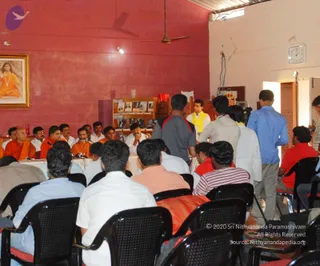 A photograph of an indoor interview or press-style gathering in a room with pink-red walls, a ceiling fan and a wall clock. On the left, a row of seated men in orange and white robes sits behind a white-draped table with microphones. A framed portrait on the left wall shows a figure in orange robes with a beaded necklace, seated in meditation. A small wooden shelf holds framed photographs. In the foreground, several people sit in black chairs with their backs to the camera, while standing men in white shirts, a man in a yellow shawl and a man in a blue shirt move around the room.