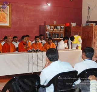 A press conference in a hall with a red-painted wall. Behind a white-draped table, several men in saffron and orange robes sit, including a bald man on the left and a man at centre with a microphone speaking. A man in a white shirt sits at the table's right end, and another man in white with a yellow shawl bows over the table. A framed portrait labelled on the wall shows a saffron-clad figure with a rudraksha mala. Microphones stand on the table, and a wooden bookshelf holds framed photographs and books. Four seated people in white shirts face the table from the foreground, one with a hand to his chin.