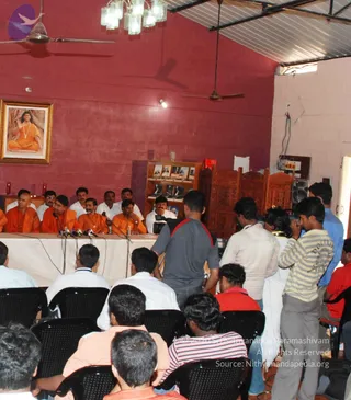 A press conference in a hall with red walls and ceiling fans. Seated at a long white-draped table are about six men; the central ones wear orange robes and are seated before microphones, while others wear white shirts. A framed portrait on the left wall shows a long-haired figure in orange seated under a tree, and a bookshelf with framed photographs stands to the right. Behind the table, a row of seated men in dark chairs and several standing people, including a man in a grey shirt with a red strap and a man in a striped shirt, watch and record the proceedings. Clusters of microphones stand on the table.