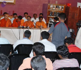 A press conference in a room with a red-painted wall, a glass-fronted bookshelf of books and framed portraits behind, and a carved wooden screen at right. Seated behind a white-draped table with several microphones on stands are men in saffron-orange robes and white shirts; the central figures wear orange robes, one with a beard and a rudraksha-style neck piece. Seated rows of people with their backs to the camera face the table. A cameraman with a video camera stands at right beside a man in a grey shirt with red trim, and a red-shirted person sits at lower right. The room has framed artwork at top left and red glass vases on a side shelf.