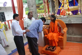 A seated figure in saffron-orange robes and a long rudraksha mala sits in an orange armchair with orange cushions on an orange carpet, before an ornate temple shrine with gold railings, a deity murti in blue and gold draped with red and orange garlands, and a kalasha. A man in a striped blue shirt and dark trousers bends forward to offer something, while a man in a white shirt stands at left and another in a pale shirt stands behind. A yellow-sashed priest in white stands at the left edge. The seated figure has long dark hair and a wide smile, with the appearance of a candidate for a person given the saffron robes, rudraksha mala and long swept hair, though the image shows the cues only partially; a person is set to unsure. A black board on the left lists text, and a red-and-white pillared structure frames the scene.