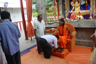 A photograph of a padapuja-style honouring inside a temple hall with orange curtains and carved stone pillars. A long-haired man in orange robes and a rudraksha mala sits on an orange cushioned chair, hands resting on the armrests, while a man in a white shirt and black trousers bends low to touch his feet placed on an orange footstool with sandals. Behind them an older man in white clothes stands, and another man in a blue striped shirt stands at left. On the altar behind, a golden throne with blue drapery and marigold garlands sits beside a small deity murti, with brass lamps and fruit offerings on the stone platform. The orange floor cloth is patterned with blue and yellow border.