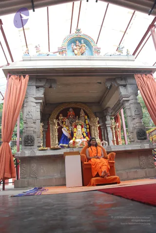 A seated man in saffron-orange robes sits in an orange armchair on a raised platform under a canopy. He has long dark hair, a rudraksha mala with a braided black cord, and his hands rest together in his lap. Behind him stands an ornate stone shrine with carved pillars, orange curtains draped at the sides, and a gilded arch framing deity murti decorated with marigold garlands. Brass oil lamps flank the shrine, and a kalasha-like vessel sits before the deities. A white lectern stands to his left and a red carpet lies on the floor.