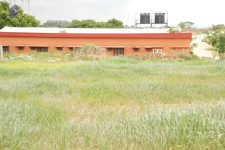 A photograph of a long single-storey building with orange walls and a red-trimmed white corrugated roof, set behind an overgrown field of tall green grass in the foreground. Two black water tanks sit on a metal stand at the right end of the roof. Rubble and dirt mounds lie along the building's front, a dirt track runs beside the right side, and trees and hazy hills appear in the background. No people are visible.