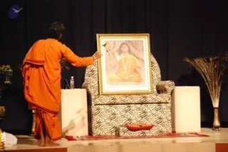 A stage event with a dark blue curtain backdrop. A man with short dark hair, wearing an orange-saffron dhoti and shawl and standing barefoot, reaches toward a large gold-framed portrait placed on a zebra-striped armchair. The portrait shows a smiling man with long dark hair, a rudraksha-style bead necklace and orange robes, seated in meditation before a tree-trunk background. Two white pedestal blocks flank the chair, a red cloth lies on the floor, a brass oil lamp stands at the left edge, and a tall vase of golden grasses stands at the right. A small glass of water sits on a side table.