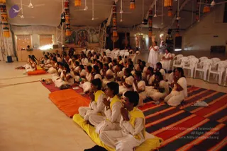 A large hall with a tent-like ceiling and hanging fabric banners in orange, yellow and red. Rows of people sit on the floor in white clothes with hands folded in prayer, facing a front area. Several devotees wear yellow shawls over white clothing and sit on yellow and orange cushions over striped rugs. A woman in white stands near the right side with her palms pressed together. A painted deity mural hangs on the back wall, and white plastic chairs line the right side of the hall. The scene suggests a satsang or meditation gathering.