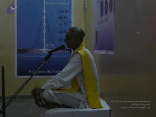A shaved-headed man in white ritual clothes and a yellow shawl sits cross-legged on a white-covered platform, speaking into a microphone on a stand. A red tilak mark is visible on his forehead. Behind him a blue poster shows a Present Moment diagram with a chart labelled 100 TPS, 80 TPS, 60 TPS, 40 TPS, 20 TPS, 0 TPS and a Time Shift strip reading Future to Past. A smaller purple poster hangs on the right wall. The room has pale yellow walls and the photo is a plain indoor view of a talk or discourse.