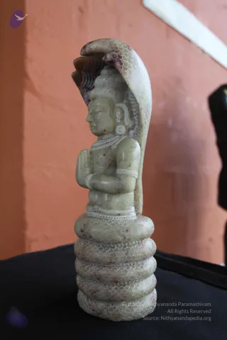 A carved pale green-grey stone sculpture of a seated deity murti sits on a black cloth against an orange-pink wall. The figure has hands folded in front of the chest and a cobra hood arching behind its head, and its base is a coiled serpent with scale patterns.