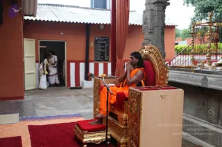 A photograph of an outdoor courtyard at a temple-like ashram. A man with long swept-back dark hair, wearing an orange saffron robe, rudraksha mala and a necklace, sits in an ornate gold throne with red cushions, hands folded in namaste, bare feet resting on a red cushioned footstool. A microphone stands before him on a wooden podium. Behind him are red and white striped walls, a corrugated roof, a gold kalasha-topped railing and a brass lamp stand. Two people in white dhoti and shawl attire stand in a doorway at left, and a red and white cloth hangs beside the throne. A printed board on the wall lists names. The seated man has distinctive long hair, saffron clothing and rudraksha adornments on a throne, consistent with the page's reference to Swamiji, so he is labelled as a probable candidate.
