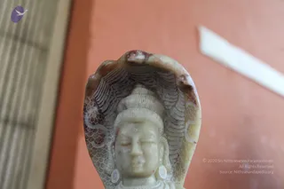 Close-up photograph of a pale carved stone statue of a deity with a cobra hood (naga) spread behind its head, showing a tiered crown-like hairstyle, carved earrings and a serene closed-eye face. The background is an orange-painted wall with a blurred wooden slatted panel at left and a white strip at upper right. No people are visible.