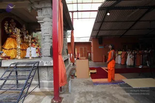A photograph of an open-air temple courtyard with a tiled roof. On the left, a shrine holds deity murti dressed in yellow and gold garments with flower garlands, with brass oil lamps in front. A standing man in saffron-orange robes faces the shrine at the centre-right, beside a wooden lectern and a red carpet. Behind him, a line of about eight people in white and saffron clothes stand with their hands folded or holding flowers. Red pillars frame the scene, and a wall with a small framed notice is visible in the background.